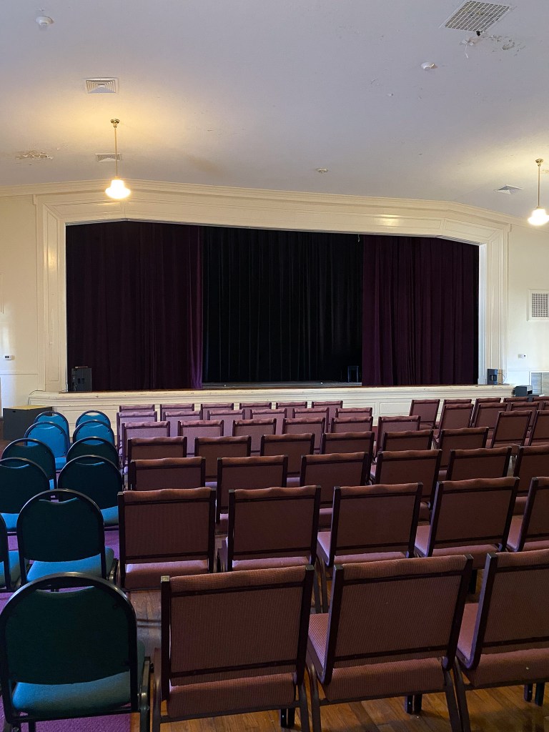 Auditorium room that includes a stage with purple curtains, white walls, and nine rows of seating