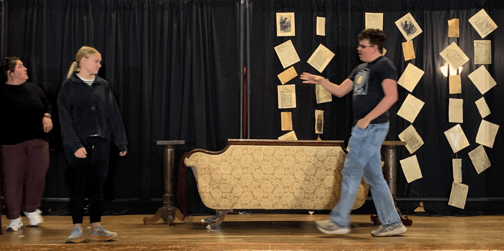 Two women and one boy on the stage at the Southside Center for the Arts rehearsing for a little women play. A gold-colored antique couch and suspended book pages sit in the background.