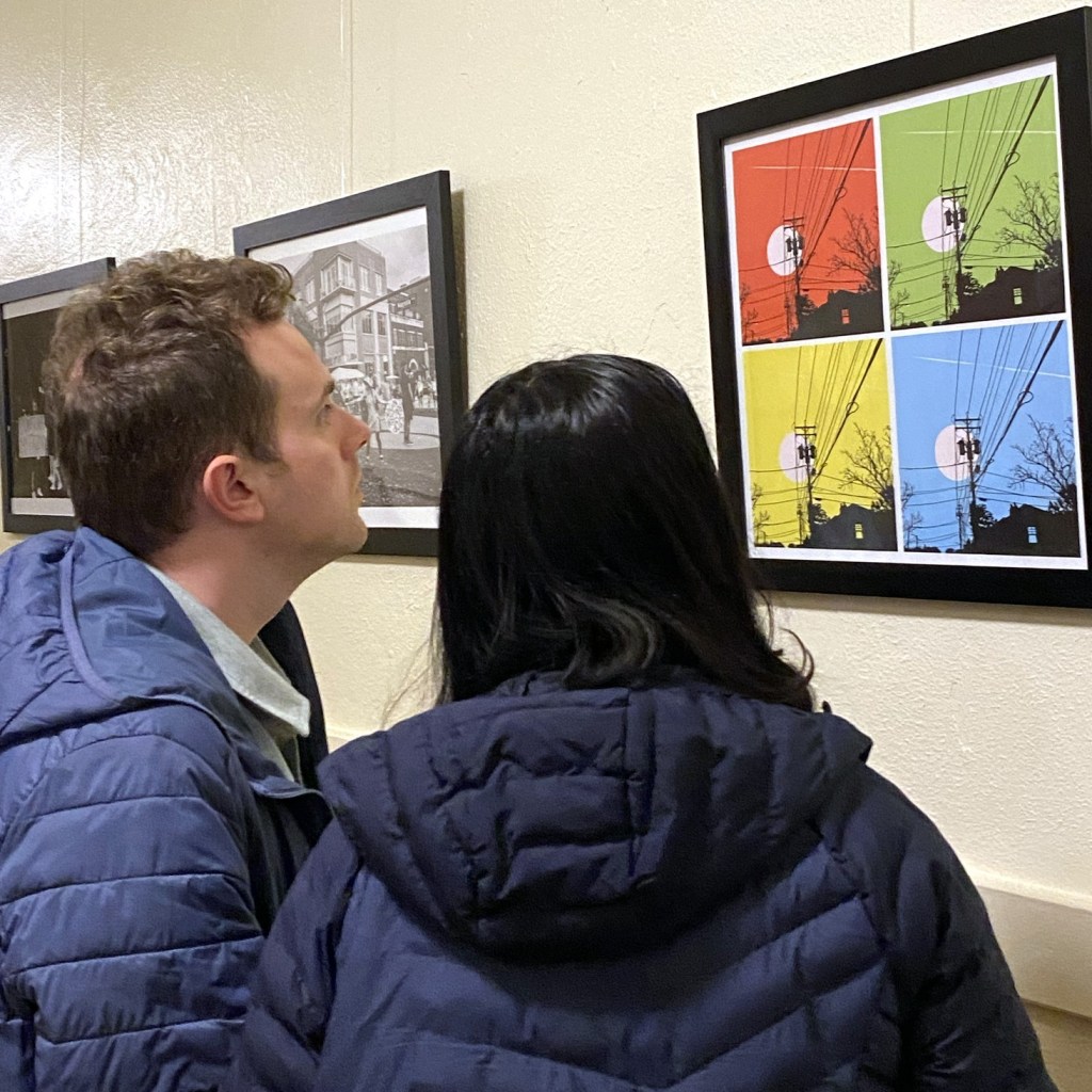 Two people looking at two-dimensional art work on the wall of the Hallway Gallery