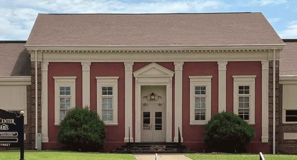 A red building with cream trim and a triangular pediment stands behind two trimmed bushes. The entrance features double doors framed by decorative columns. A black sign to the left reads ‘Center for the Arts’ with additional smaller text below.