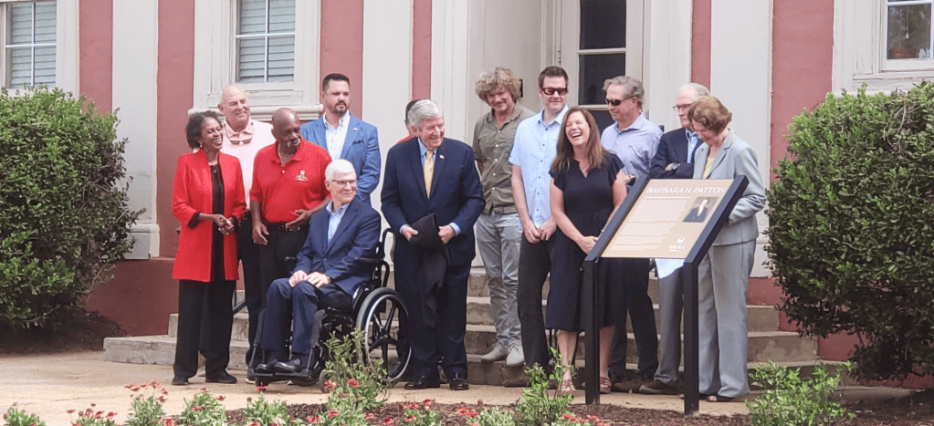 A group of people stand outdoors in front of a red‑and‑white building during a plaque‑unveiling event. One person is seated in a wheelchair, and a commemorative plaque featuring the name ‘Barbara H. Patton’ is displayed on a stand in front of the group. Trimmed bushes and flowering plants are visible in the foreground.