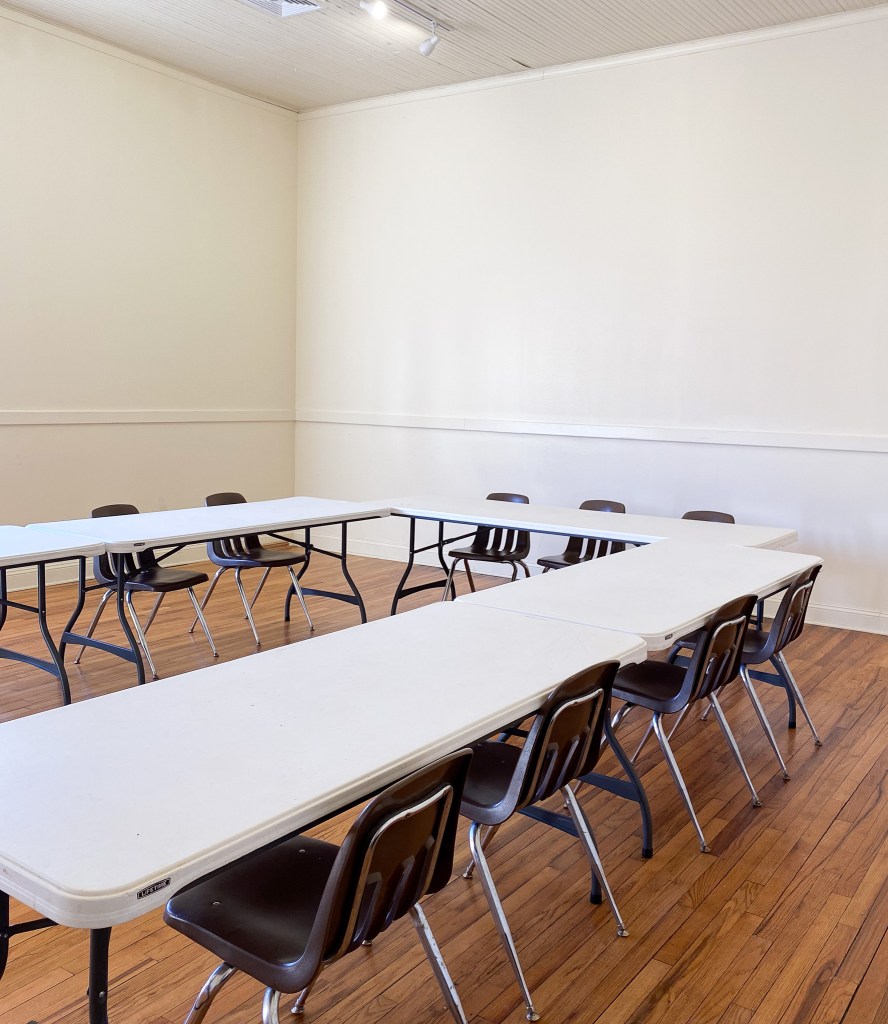 A small classroom with white tables in a U formation and brown chairs around the tables set up for a meeting scenario.