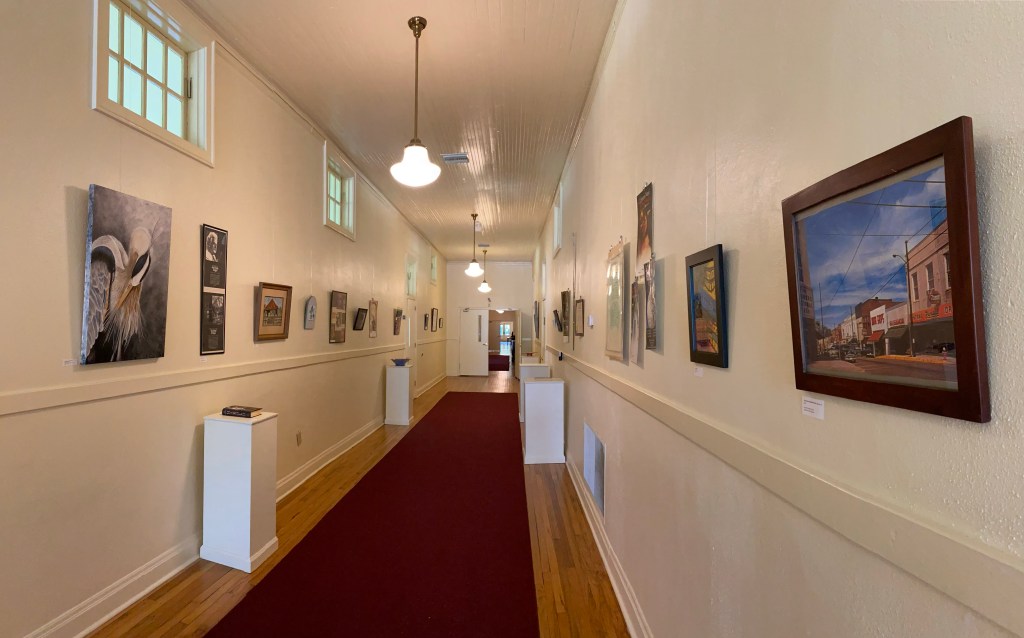 Long hallway gallery with framed artwork on cream walls, red carpet runner, warm ceiling lights, and pedestals displaying small pieces.